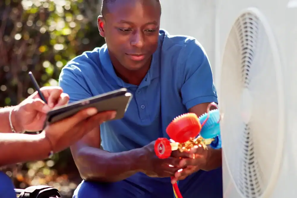 A technician in a blue shirt checks air conditioning unit gauges while an HVAC Contractor Essex County,NJ takes notes on a clipboard. They are working outdoors with sunlight in the background.