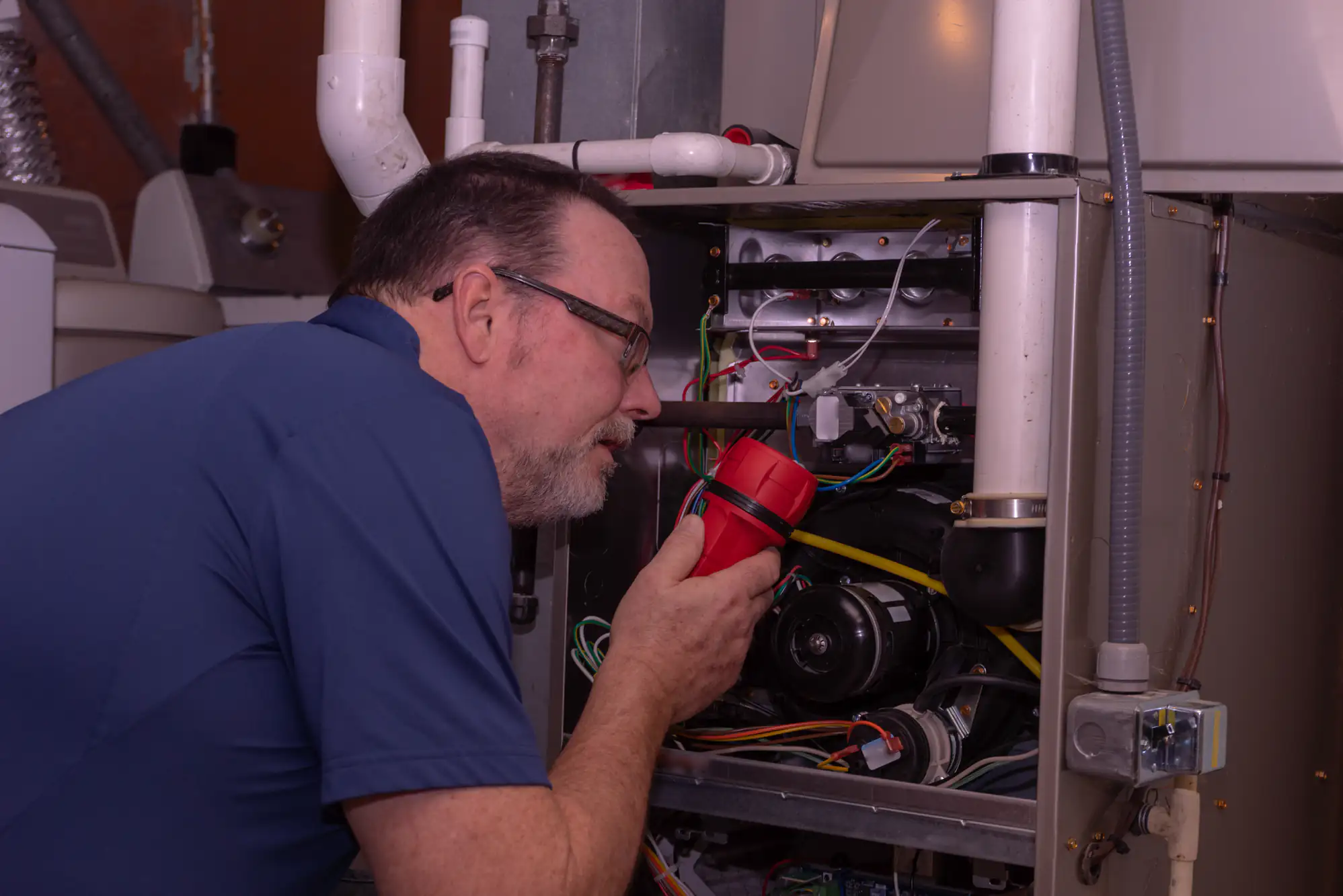 A man wearing glasses and a blue shirt inspects the inside of a furnace, holding a red flashlight and looking closely at the wiring and components.