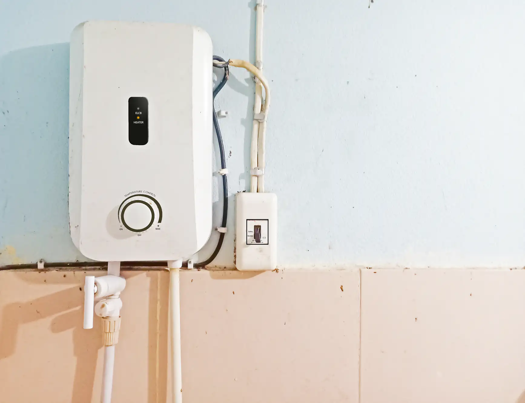 A white electric water heater mounted on a wall above beige tiles, with visible pipes, wires, and a switch next to it on a light blue painted wall.
