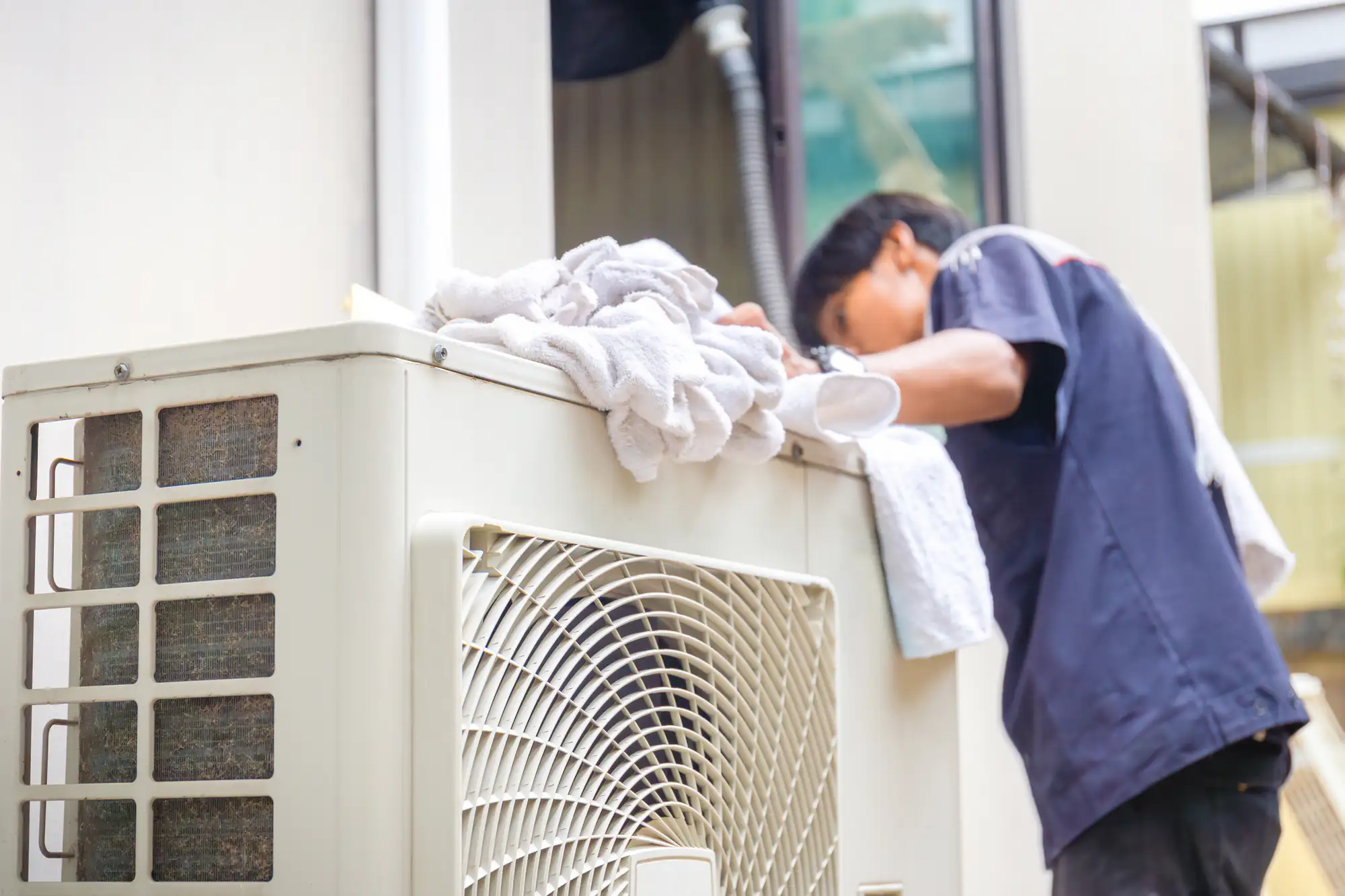 A technician repairs an outdoor air conditioning unit, with towels placed on top for protection during work.