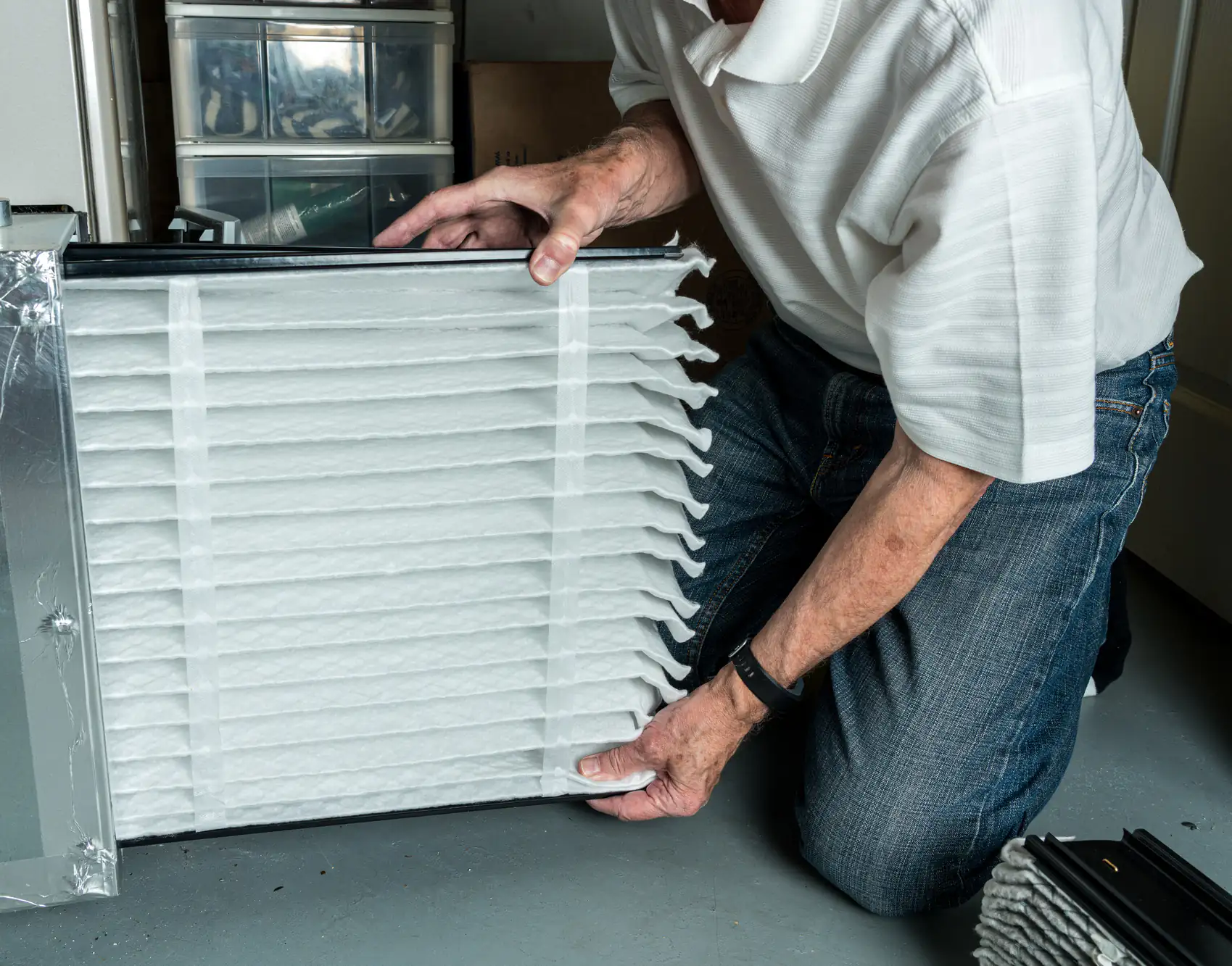 A person installs a large, pleated air filter into a home HVAC unit positioned on a gray floor.