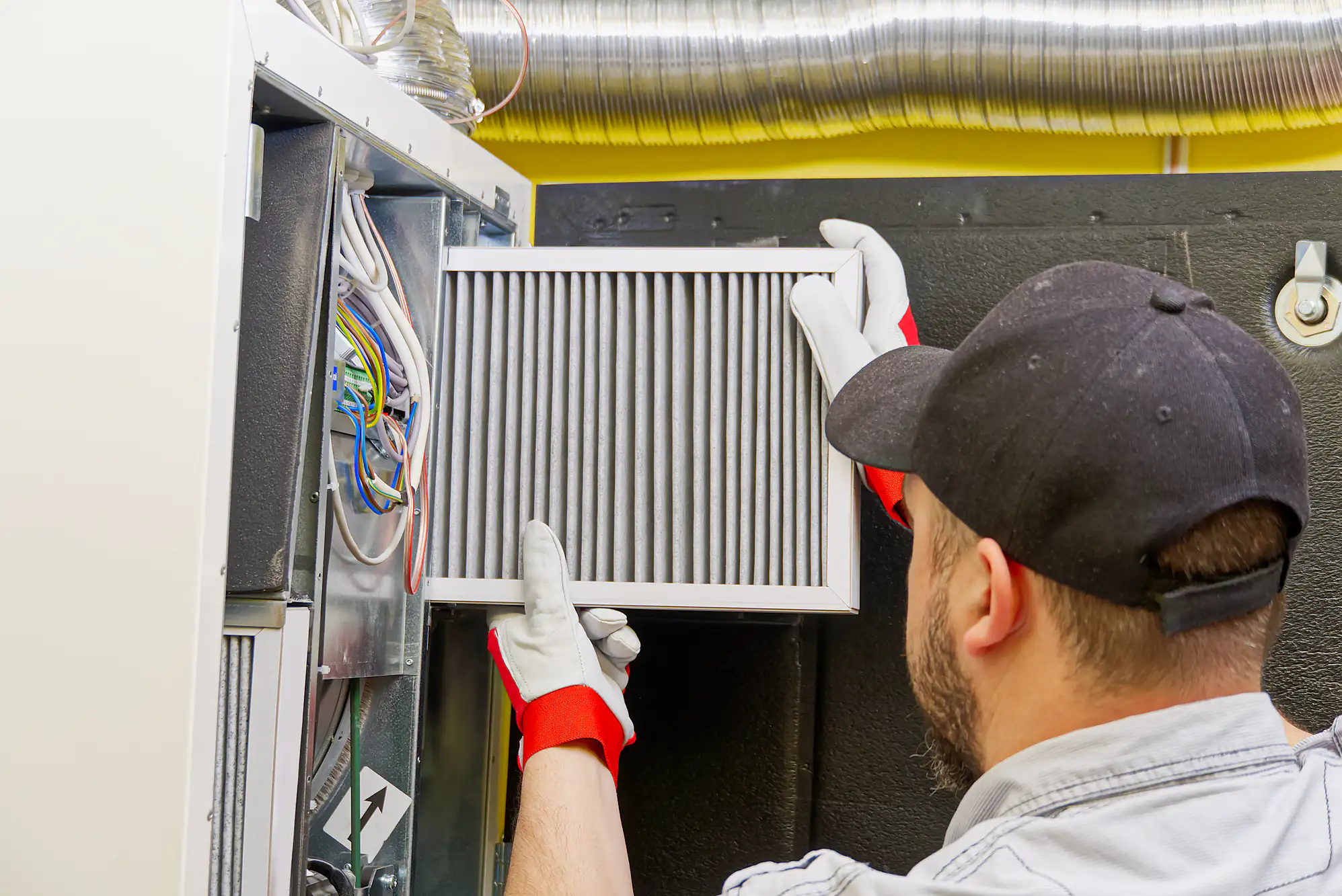 A worker wearing gloves replaces an HVAC air filter near exposed ducts and wiring for better air quality.