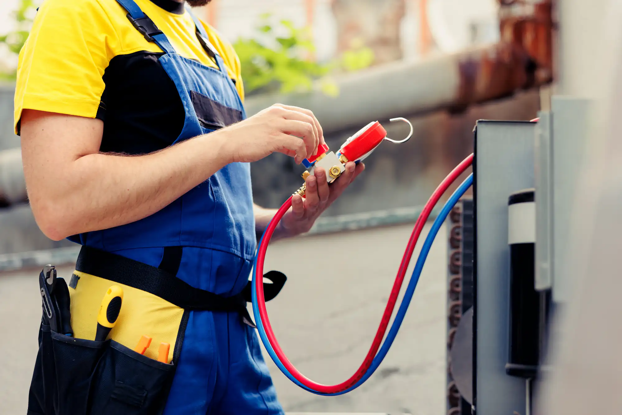 A technician in blue overalls checks HVAC gauges while servicing an outdoor air conditioning unit.