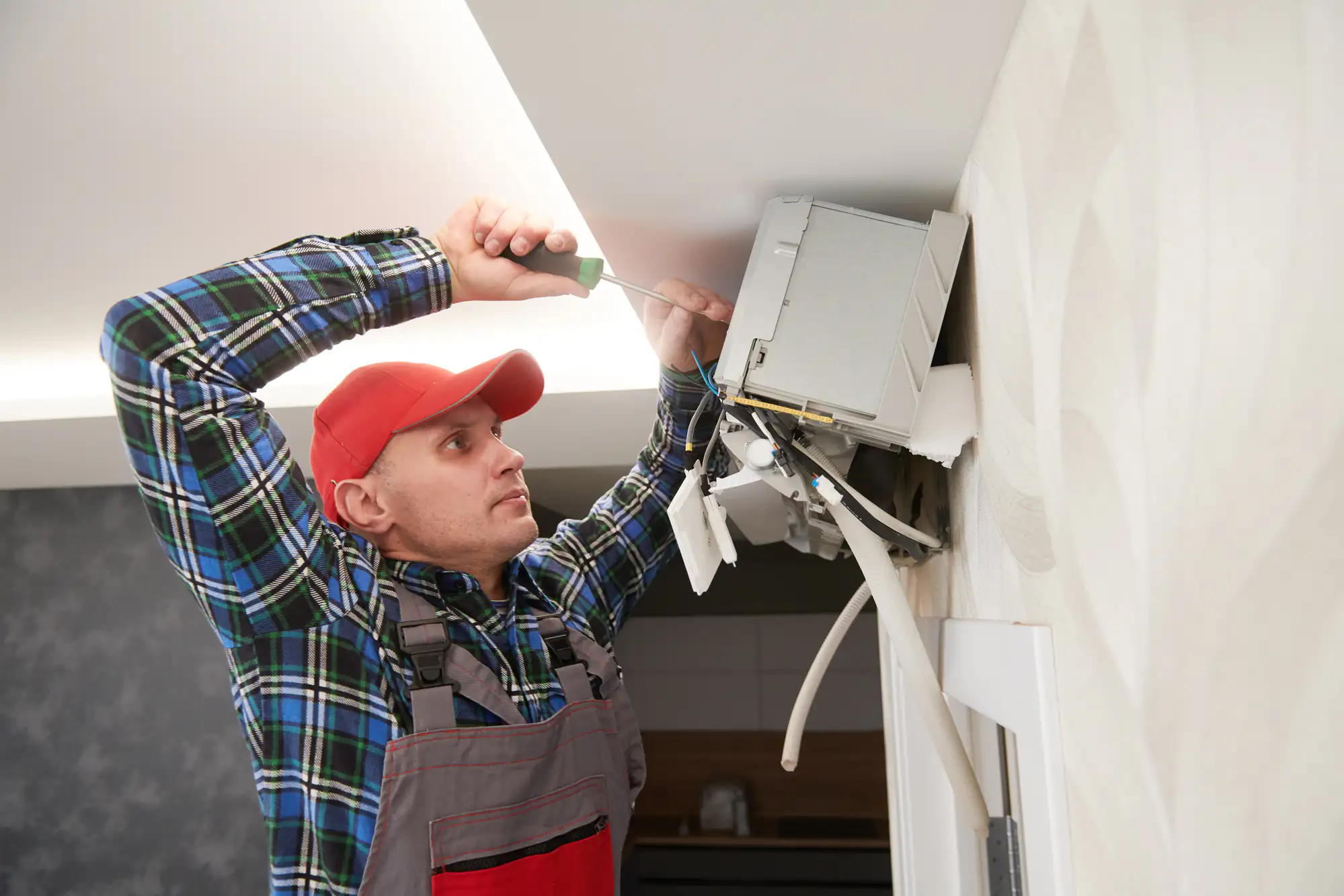 Technician wearing a red cap uses a screwdriver to repair a wall-mounted air conditioning unit.